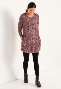Eine lächelnde Frau schaut nach unten, trägt eine weinrote gemusterte Tunika mit Taschen, schwarze Leggings und schwarze Knöchelstiefel und steht vor einem weißen Hintergrund.