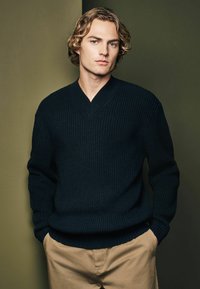 Marineblå ribstrikket sweater med V-hals, lange ærmer og en tyk struktur, parret med beige bukser. Baggrunden har dæmpede grønne nuancer.
