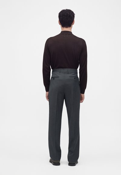 Un homme est debout, tourné de dos, portant une chemise sombre à manches longues rentrée dans un pantalon de costume gris taille haute avec des chaussures noires.
