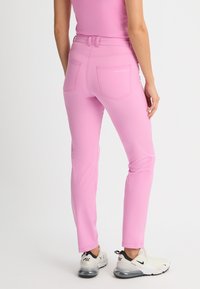 Pink fitted pants with a smooth texture, five-pocket design, and contrasting stitching detail, paired with white and black sneakers.