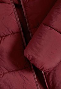 Burgundy puffjacka med en glänsande yta, som har en dragkedja fram och sydda sektioner för isolering. Ärmen är något invikt.