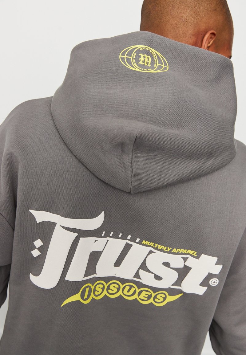 Sweat à capuche gris avec un grand texte graphique blanc et jaune au dos. Comprend un petit logo jaune imprimé sur la capuche. Texture douce.