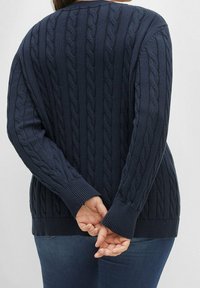 Osoba nosząca granatowy sweter w warkocze, z rękami splecionymi za plecami, w połączeniu z ciemnoniebieskimi dżinsami, pokazana od tyłu.