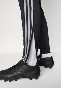 Black athletic pants with grey side stripes feature zippers along the legs, paired with black soccer cleats and white socks.