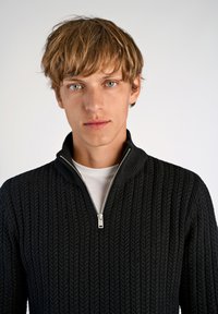 Schwarzer Strickpullover mit strukturiertem Muster, Reißverschlusskragen und einem weißen Unterhemd. Der Stoff erscheint weich und hat eine taillierte Passform.