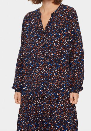 Kvinde iført langærmet sort bluse med blåt, hvidt og brunt abstrakt prikmønster og matchende nederdel mod hvid baggrund.