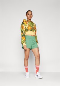 Trumpas hoodie su geltonais ir žaliais gėlėtais raštais, derinamas su žaliais šortais. Balti sportbačiai ir oranžiniai dryžuoti kojinės užbaigia aprangą.