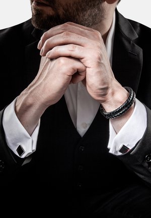 Black suit with a white dress shirt. Features metal cufflinks, a braided black bracelet, and clasped hands with visible veins.