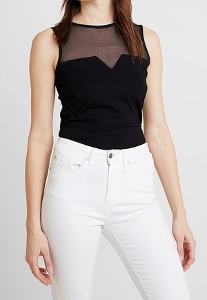 Kobieta ubrana w czarny top bez rękawów z przezroczystym dekoltem i białe, wysokowalne dżinsy, stojąca na jednolitym tle.