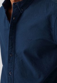 Granatowa koszula z guzikami wykonana z bawełny o miękkiej fakturze. Posiada ostry kołnierzyk, guziki z przodu oraz małe logo na piersi.