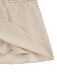 Svetlo bež krilo iz teksturirane tkanine z majhnimi dvignjenimi pikami, z elastičnim pasom in slojevitim robom za dodatne podrobnosti.