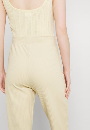 Vrouw draagt een lichtgele geribde mouwloze top en hooggetailleerde geribde broek, gezien van achteren tegen een effen achtergrond.
