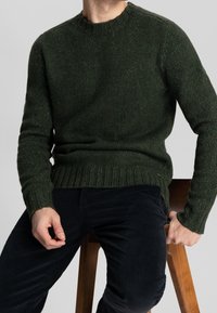 Dunkelgrüner Strickpullover mit Rundhalsausschnitt sowie gerippten Bündchen und Saum. Kombiniert mit dunklen Cordhosen, sitzt auf einem Holzschemel.