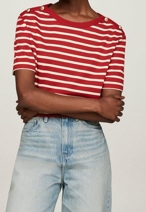 Personne portant un haut à manches courtes rouge et blanc à rayures horizontales avec des boutons dorés sur les épaules et un jean bleu clair taille haute, bras croisés.