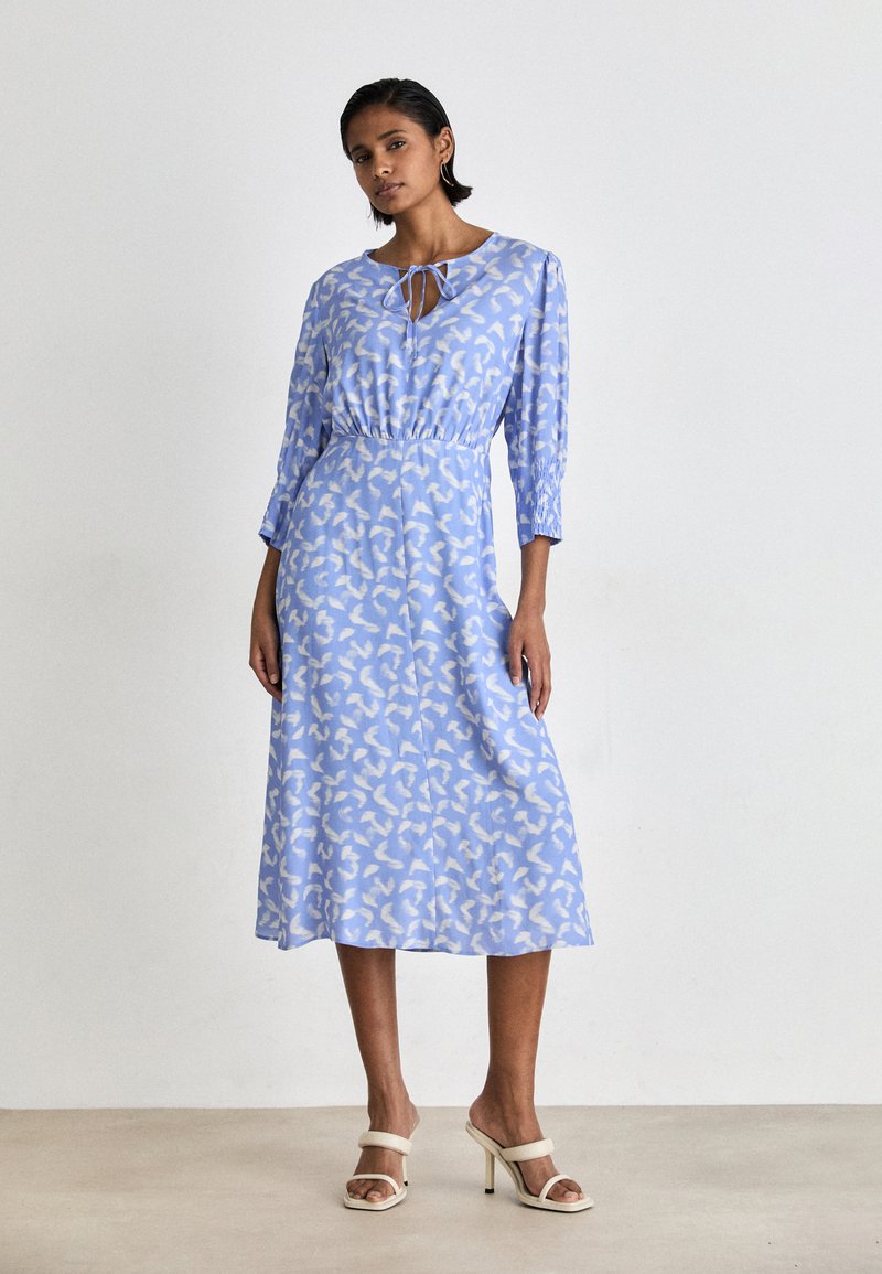 Rochie midi albastră cu un model abstract alb, mâneci lungi, guler legat și talie elastică. Țesătură netedă și siluetă fluidă.