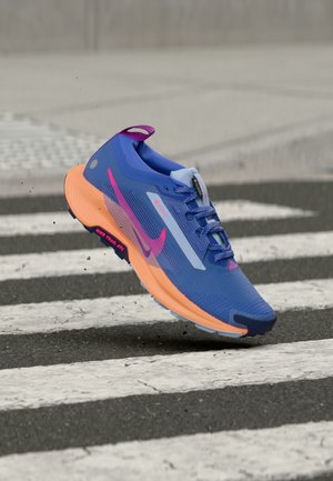 Mėlynas ir oranžinis Nike bėgimo batelis su rožine svastika, ore virš pėsčiųjų perėjos ant asfalto, šalia pado matomi purvo dalelės.