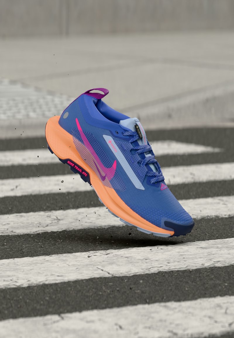 Μπλε και πορτοκαλί παπούτσι για τρέξιμο της Nike με ροζ λογότυπο, στον αέρα πάνω από διάβαση πεζών σε άσφαλτο, με σωματίδια βρωμιάς κοντά στη σόλα.