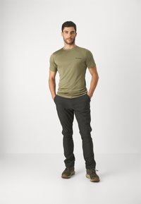 Camisa verde oliva de manga corta con un diseño ajustado, que presenta un logo. Combinada con pantalones negros y robustos zapatos de senderismo marrones.
