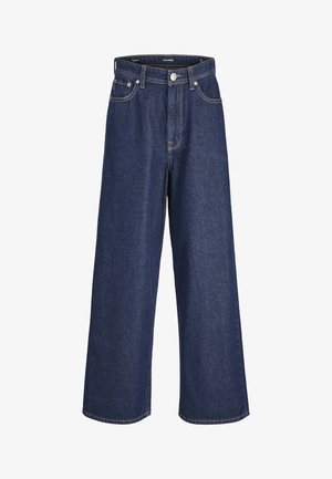 Un enfant sourit tout en portant un t-shirt blanc, une écharpe bleue, une casquette marine et un pantalon bleu ample, se tenant devant un fond uni.