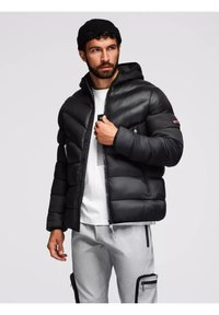 Giacca a vento nera con cappuccio, design trapuntato, zip frontale e tasche laterali. Indossata sopra una maglietta bianca, abbinata a pantaloni grigi chiari.