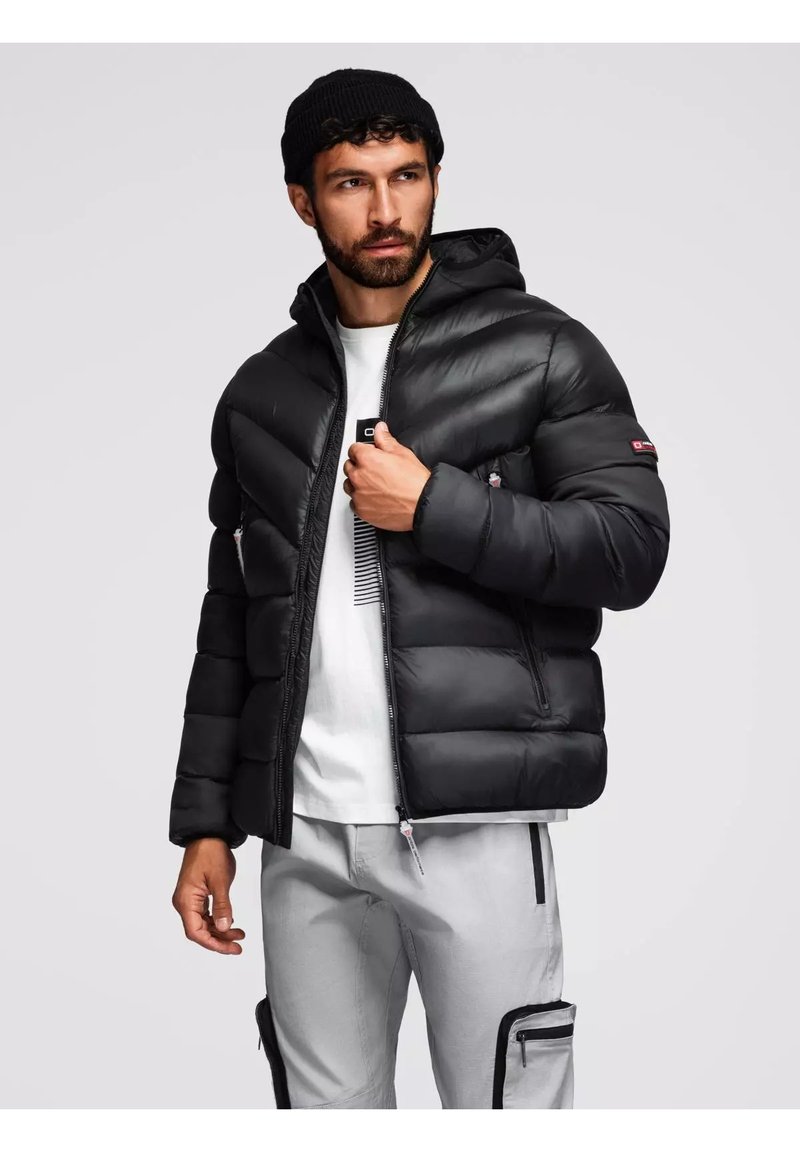 Giacca a vento nera con cappuccio, design trapuntato, zip frontale e tasche laterali. Indossata sopra una maglietta bianca, abbinata a pantaloni grigi chiari.