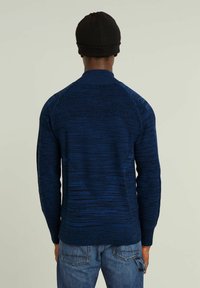 Maglione lavorato a maglia di colore blu scuro con un motivo testurizzato. Presenta un colletto alto e polsini a costine, indossato con jeans in denim blu.