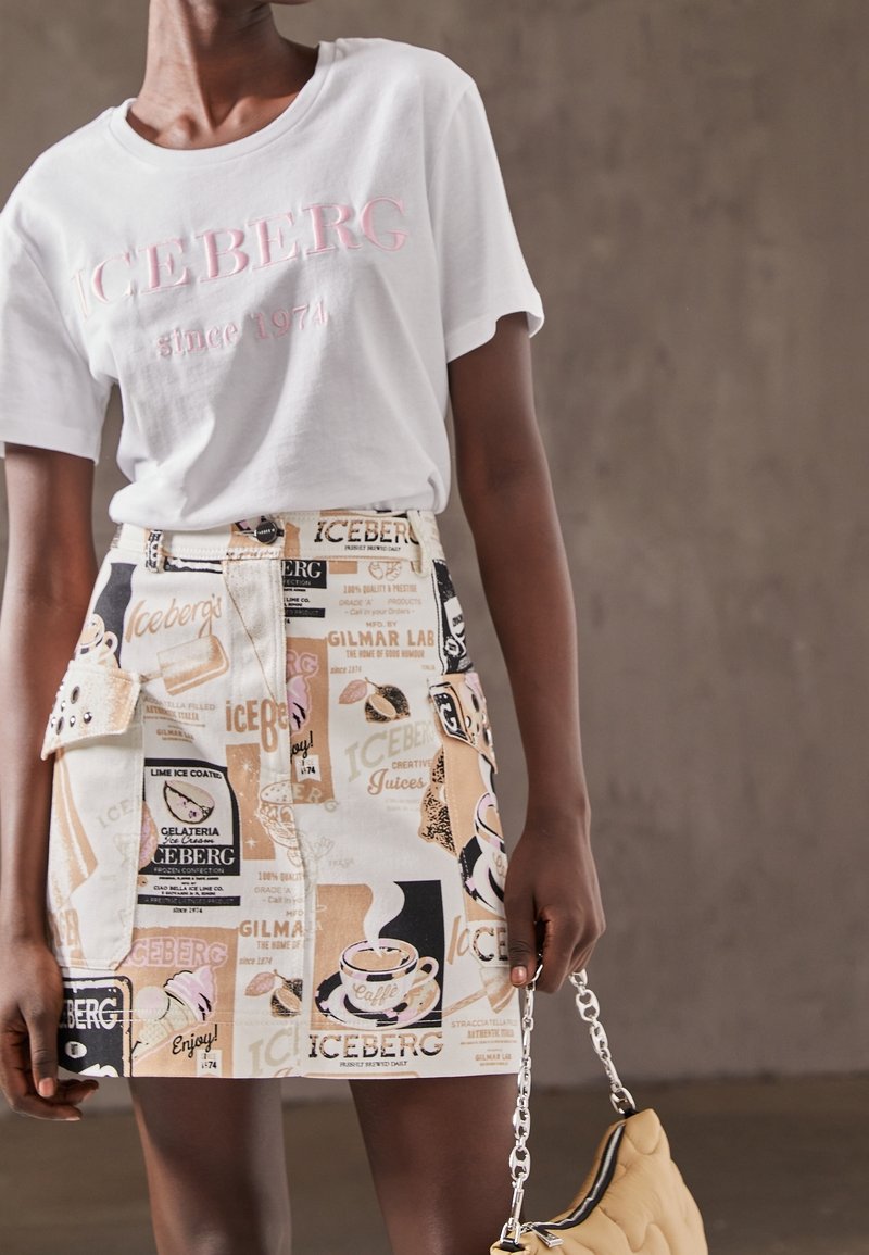 Personne portant un T-shirt blanc "ICEBERG depuis 1974" et une jupe imprimée avec des patchs texte et graphiques, tenant un sac matelassé beige.