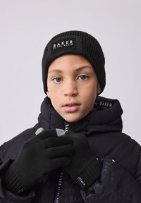 Black ribbed knit beanie with a logo patch, paired with black gloves featuring gray tips. Warm, textured fabrics; hooded black jacket in background.