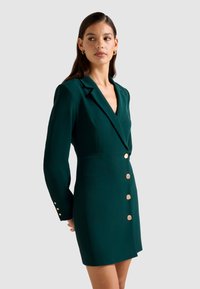 Donna che indossa un vestito blazer verde scuro con bottoni d'oro, maniche lunghe e orecchini a cerchio dorati, che guarda di lato su uno sfondo neutro.