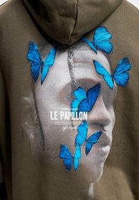 Olivazöld kapucnis pulóver, amelyen egy arc grafika és élénk kék pillangó nyomatok találhatók. A grafika alatt a szöveg: "LE PAPILLON." Puha anyagú textúra.