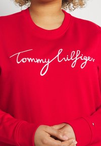 Czerwona bluza z miękkiego materiału z białym, kursywnym logo "Tommy Hilfiger". Rybosz w okolicy szyi i mankietów, z małym akcentem logo na rękawie.