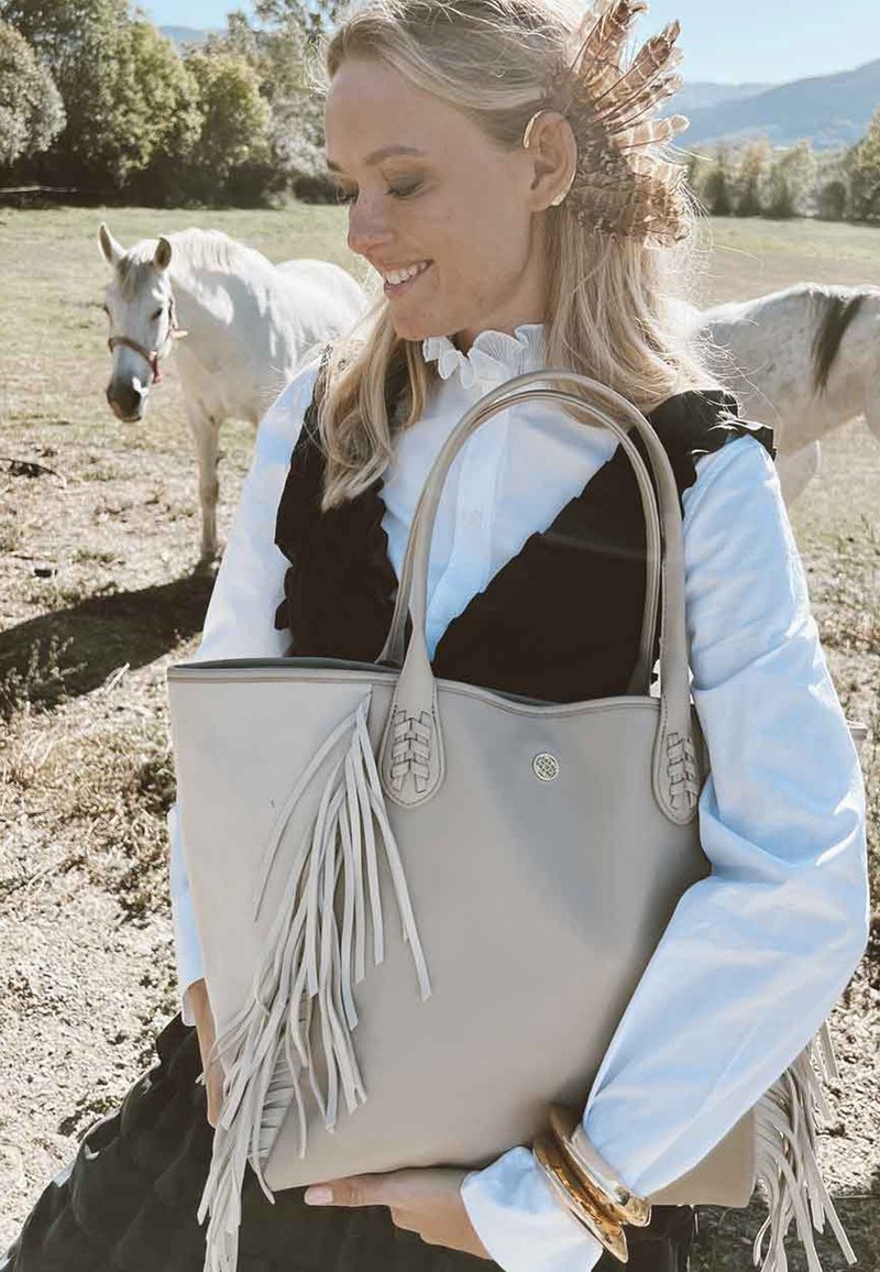 Sac fourre-tout en cuir taupe avec des accents tissés, des franges et deux poignées robustes. Le sac a une texture lisse et un emblème de logo rond.