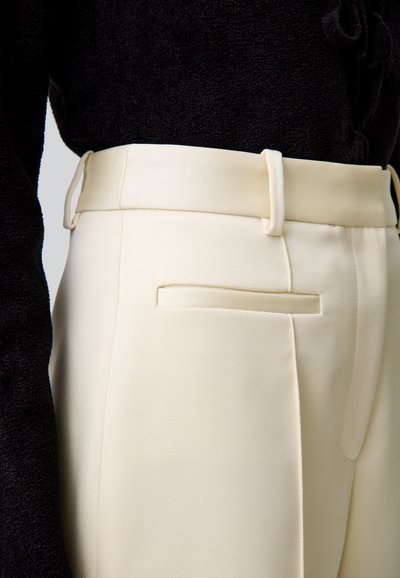 Un pantalon couleur crème en tissu texturé, avec une coupe slim, un devant plat et une poche arrière unique. Des passants de ceinture soulignent la taille.