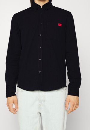 Man draagt een zwart overhemd met knopen en een rode logo patch op het borstzakje, gecombineerd met lichte spijkerbroek, voor een effen achtergrond.