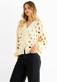Cream knit cardigan with a loose, open design, featuring a deep V-neck, large buttons, and textured patterns. Worn over dark jeans.