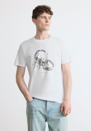 Jeune homme aux cheveux bouclés portant un t-shirt blanc avec une illustration détaillée d'un scorpion noir et un jean bleu clair, debout devant un fond uni.
