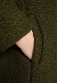 Tessuto di lana verde scuro, strutturato, con una finitura morbida e pelosa. Presenta una tasca laterale, sottilmente integrata nel design.