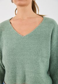 Pull vert clair en maille avec un col en V, présentant une texture côtelée et une coupe courte. Tissu doux et ajustement légèrement ample au niveau des épaules.