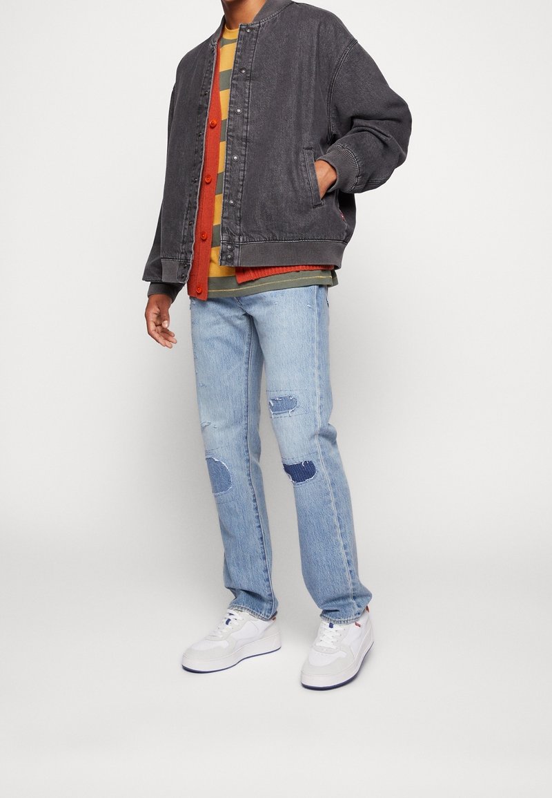 Personne portant une veste bomber noire sur un cardigan orange et une chemise à rayures, un jean bleu clair avec des patchs et des baskets blanches.