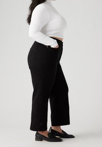 Jeans negros de pierna recta con acentos de botones de cobre, combinados con un cuello de tortuga blanco de canalé. Una textura suave y zapatos negros pulidos completan el look.