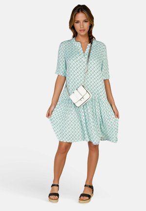 Kvinde iført en lyseblå mønstret kjole med albuelange ærmer, sorte platåsandaler og en hvid crossbody-taske, der står mod en ensfarvet baggrund.