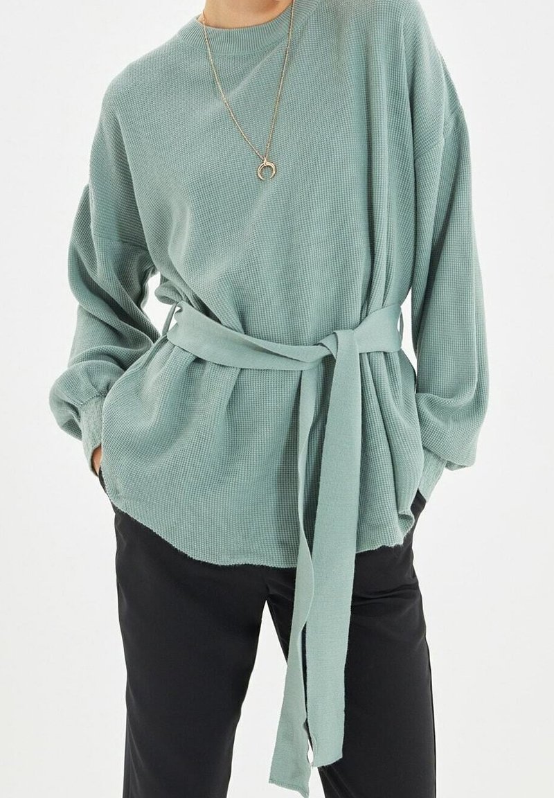 Femme portant un haut à manches longues vert clair texturé avec une ceinture assortie nouée à la taille et un pantalon noir, les mains dans les poches.