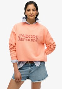 Sudadera melocotón con "J'ADORE SUPERDRY" impreso en rojo oscuro, superpuesta a una camisa de cuello a rayas azul y blanca, combinada con pantalones cortos de denim.