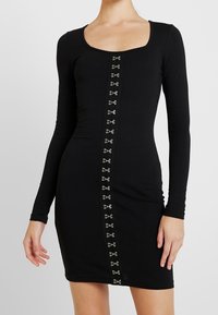 Černé dlouhé šaty s obdélníkovým výstřihem, v přední části mají středový panel zdobený stříbrnými kovovými detaily.