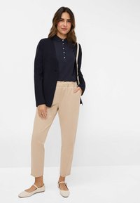 Marineblauwe polo met knoopdetails, een getailleerde zwarte blazer, beige taps toelopende broek en lichtgekleurde ballerina's met een bandje. Eenvoudige en casual outfit.