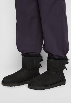Black suede ankle boots with fur lining paired with loose dark pants, shown from mid-calf to feet on a light background.