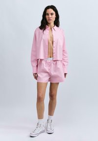 Helles rosa, kurz geschnittenes Hemd mit Knöpfen, langen Ärmeln und einer Brusttasche, kombiniert mit passenden Shorts. Trägt weiße Sneaker und Socken.