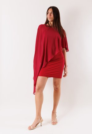 Donna in piedi che indossa un vestito corto rosso con una manica a mantella asimmetrica svolazzante, abbinato a sandali beige con tacco alto, su sfondo bianco.