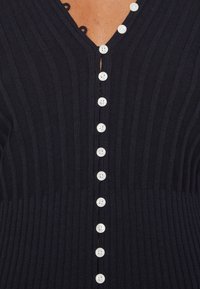 Cardigan a coste di un blu navy scuro, con scollatura a V, una fila di grandi bottoni bianchi sul davanti e anelli decorativi per i bottoni.