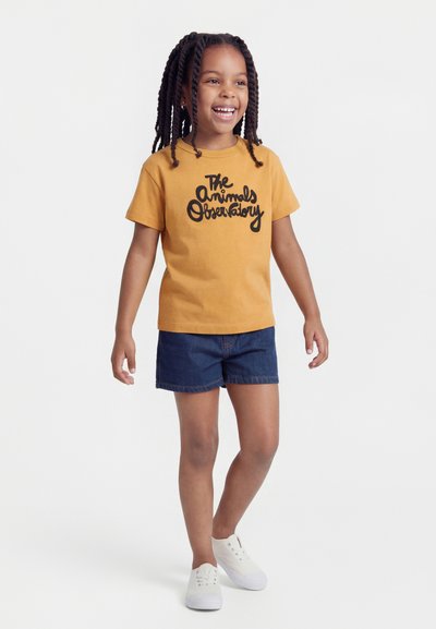 Ragazza sorridente con capelli intrecciati che indossa una t-shirt giallo senape con la scritta "The Animals Observatory", shorts di jeans e sneakers bianche.